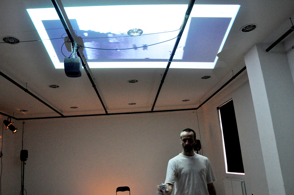 The installation space with ceiling video projection, quadraphonic loudspeakers, and a performer holding a controller.